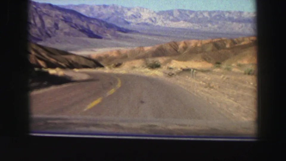 1969 home-movie frame showing a car traveling along a curving mountain road in dry California terrain.