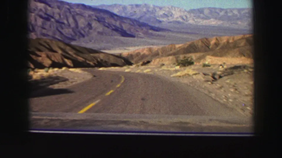 Car-window view of a winding road through rugged dry hills, with distant mountains in a 1969 8mm travel clip.