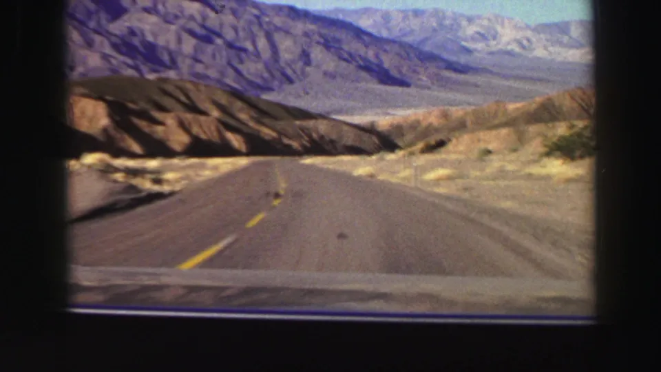 A car approaches a bend on a rural two-lane road through dry hills and distant mountain ranges in 1969 8mm footage.