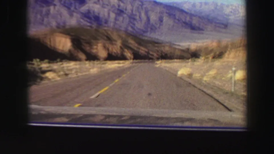 A blurred 8mm car-travel frame shows a rural road, yellow line, dry hillside, and small roadside marker in mountain country.