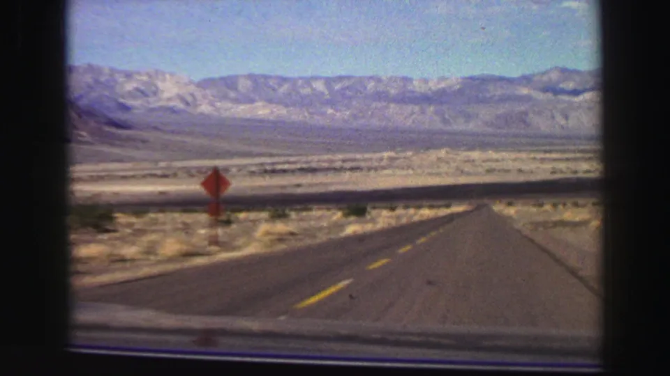 A 1969 8mm road-travel view shows a two-lane road, red roadside sign, open valley floor, and distant mountain range.