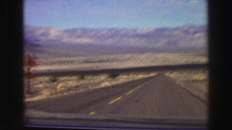 1969 8mm road-trip view from a vehicle on a desert highway toward distant California mountains.