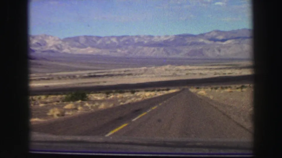 Vintage 8mm frame showing a road through sparse desert toward distant mountains in California travel footage.