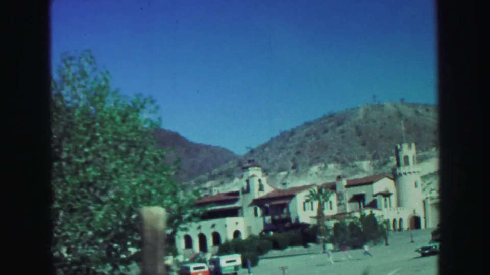 The 1969 home-movie view shows a pale building with arches and tower details, roadside trees, parked cars, and small distant visitors.