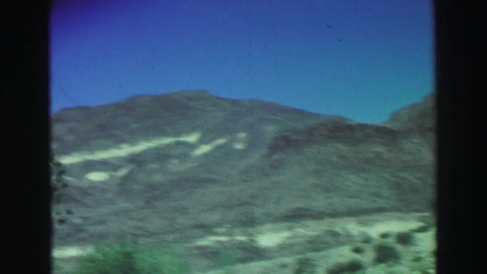 Lake Tahoe mountain hillside filmed from a moving vehicle, with pale dry slopes, scrub vegetation, and blue sky in a soft 8mm frame.
