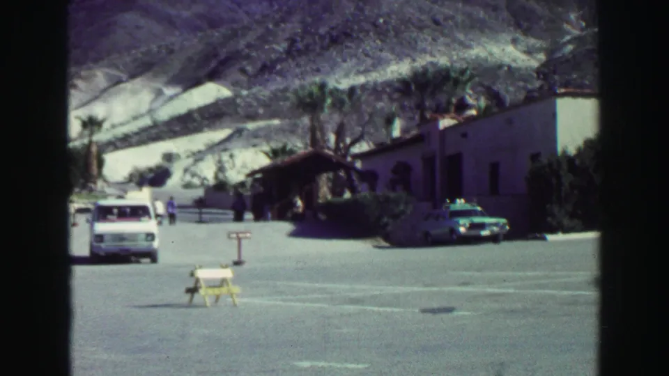 Lake Tahoe church parking area in 1969 with a white van, police car, small distant visitors, palm-like trees, and mountain slopes behind.
