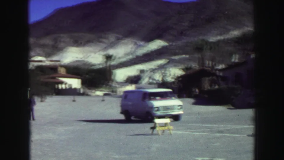 White van drives across a Lake Tahoe tourist parking area with low buildings and a mountain slope in the distance.