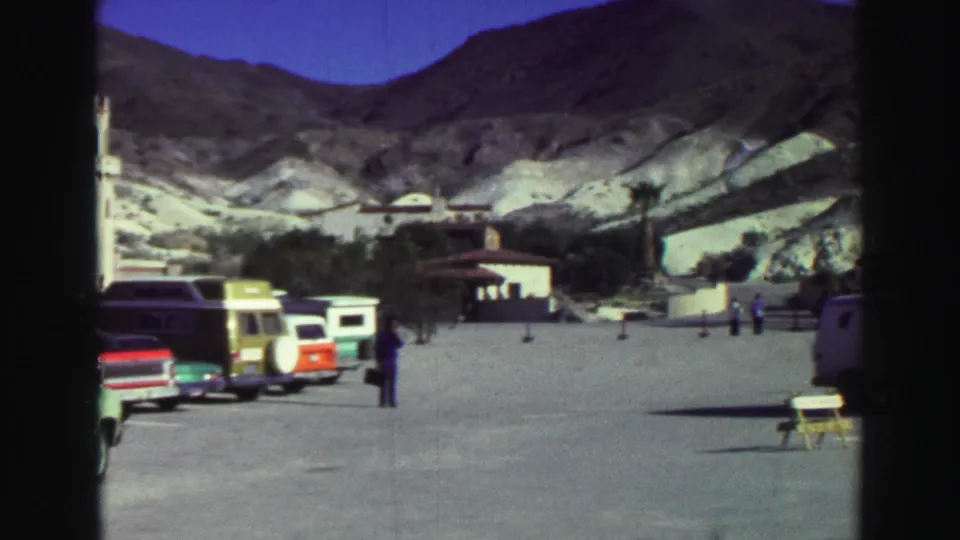 Lake Tahoe parking area with parked 1960s vehicles, scattered tourists, low buildings, and pale mountain slopes.