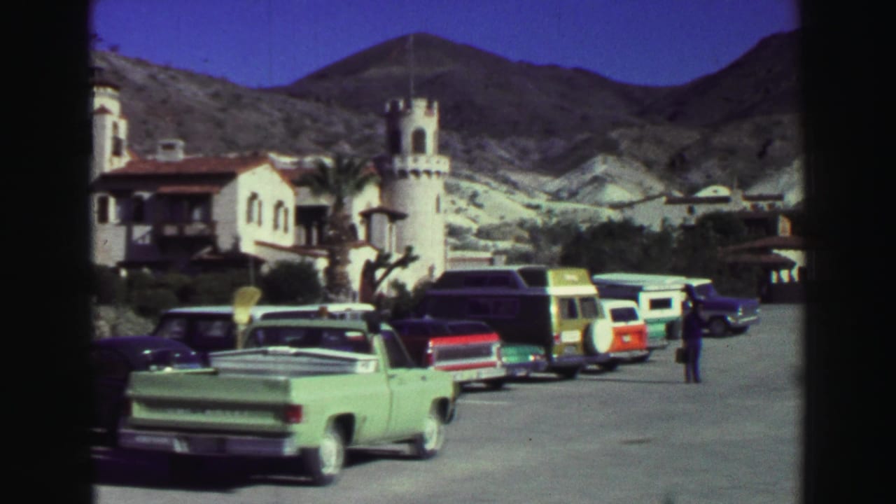 Classic cars parked before a white Spanish-style resort building with mountains behind.