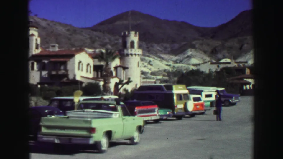 1969 Lake Tahoe view of parked cars, including a green pickup and red station wagon, outside a white resort building below rugged hills.