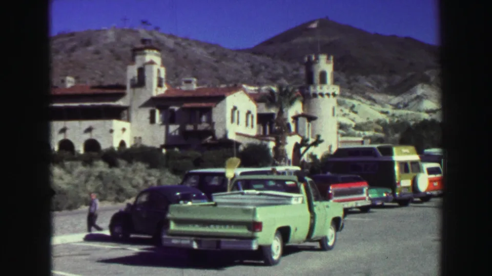 1969 Lake Tahoe resort parking view with a green pickup, dark car, vans, pedestrians, and a white Spanish-style building against hills.