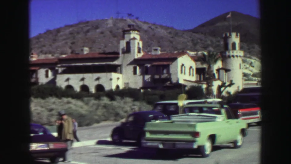 1969 Lake Tahoe home-movie frame showing a white Spanish-style resort facade, parked classic cars, pedestrians, shrubs, and mountain slopes.