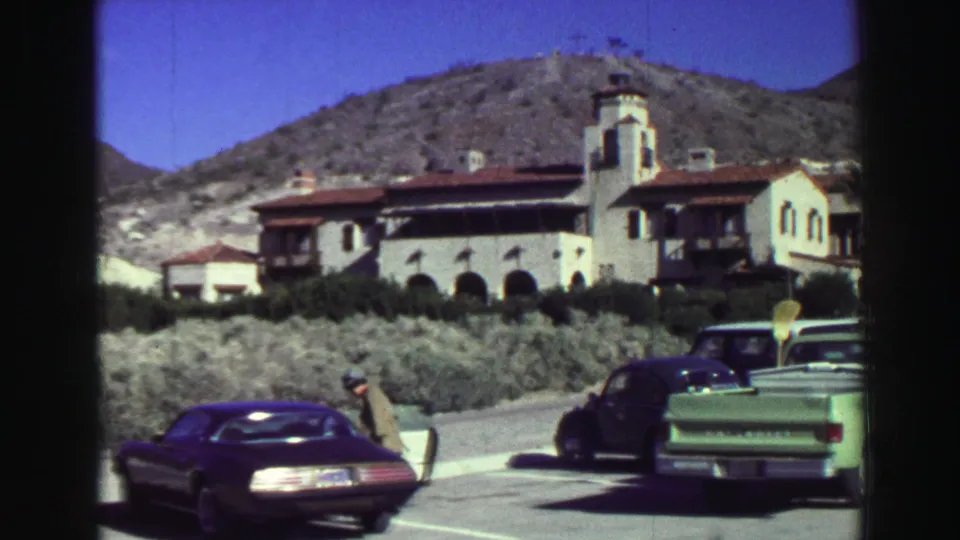 1969 Lake Tahoe parking-lot view with a white resort building, green pickup, dark coupe, pedestrians, shrubs, and hills in bright sun.