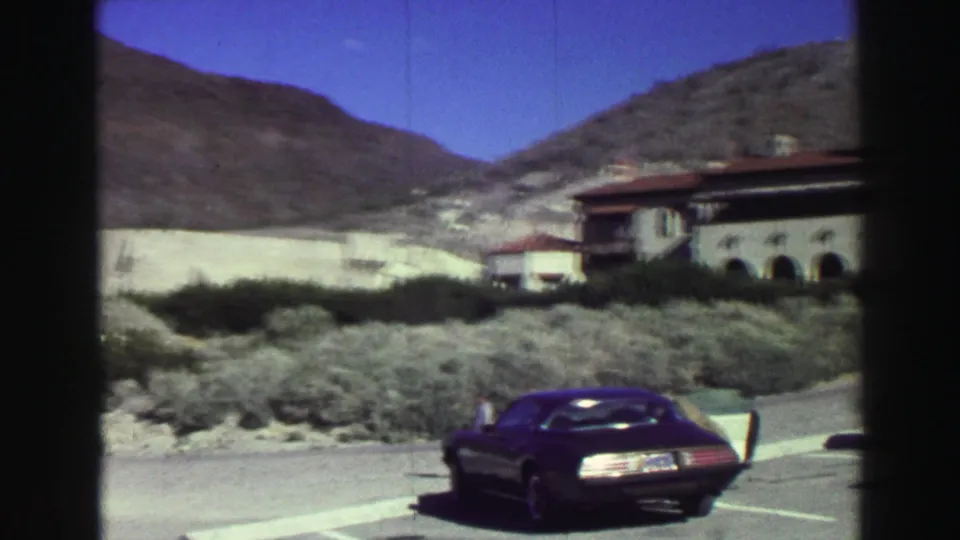 1969 Lake Tahoe frame with a dark classic car in the foreground, a stucco building at right, and a dry mountain slope behind it.