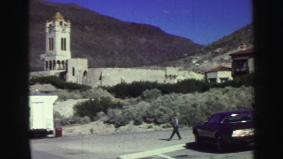 1969 Lake Tahoe home-movie frame showing a bell tower, dry brush, small white structure, parked car, and a man walking across the foreground.