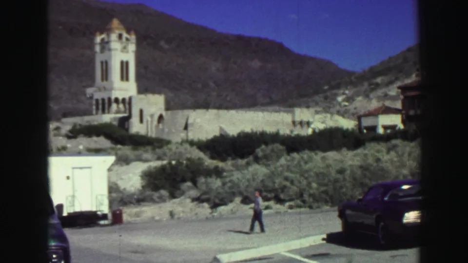 1969 Lake Tahoe frame with the bell tower at left, hillside brush behind, a walking man near center, and a dark car at the right edge.