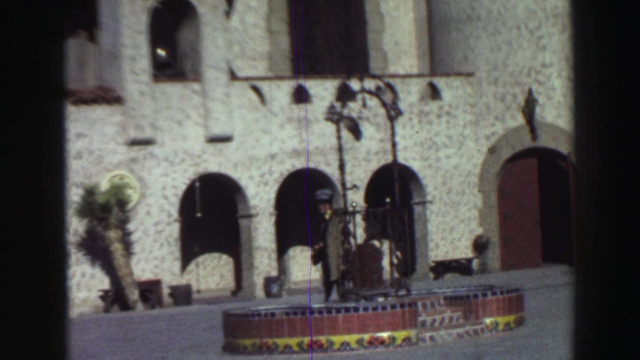 Stucco building with arched openings, fountain, lamp post, and a man near the entrance.