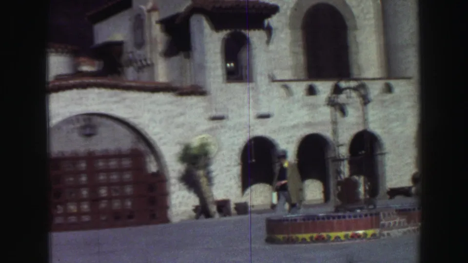 1969 Lake Tahoe frame with a man passing a Spanish Colonial Revival-style facade, red door, arches, and fountain.