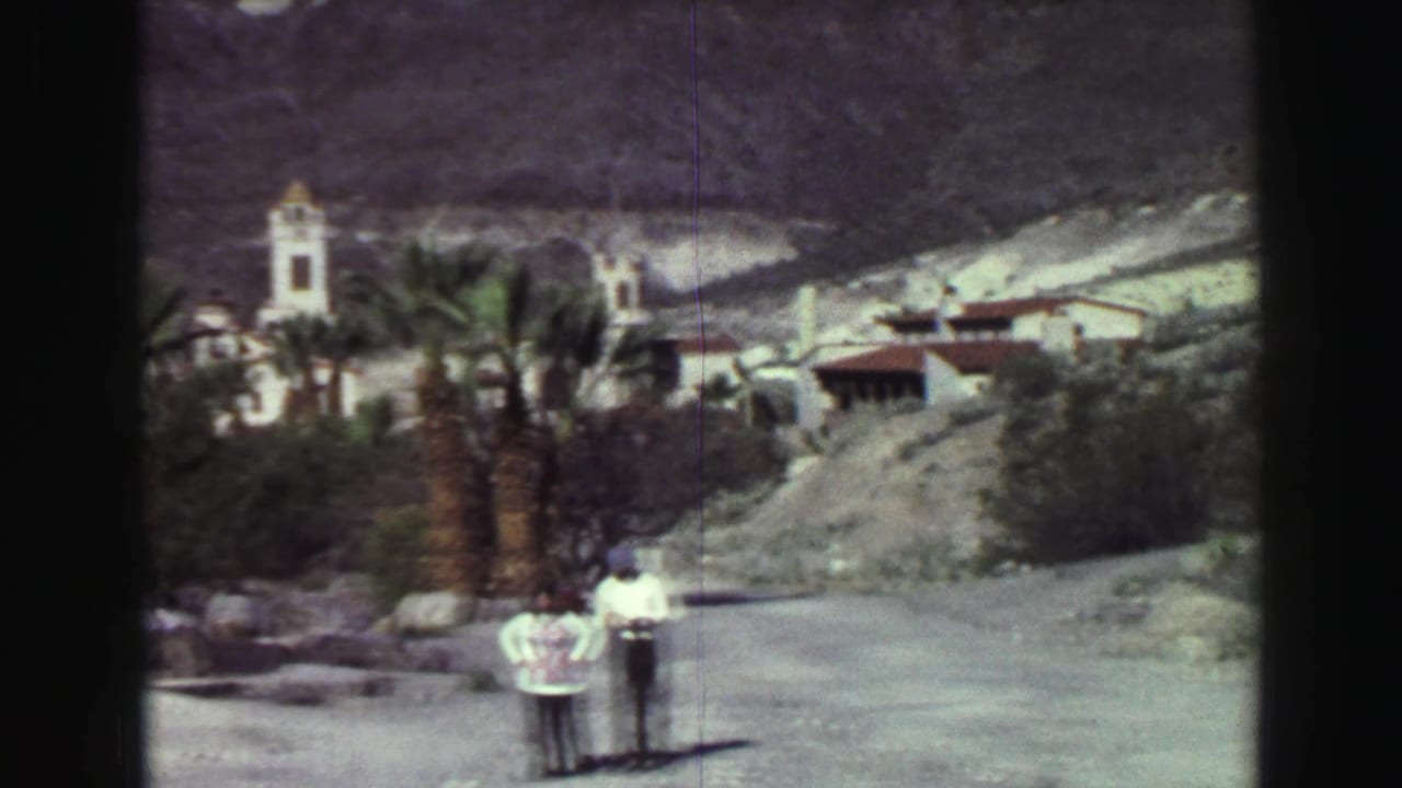 Two people stand on a gravel path with palms, rocks, buildings and mountains behind them.