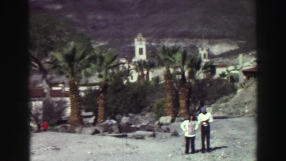 1969 8mm frame from the Lake Tahoe metadata with two visitors in bright sun near palms, rocks, low structures and a white tower.