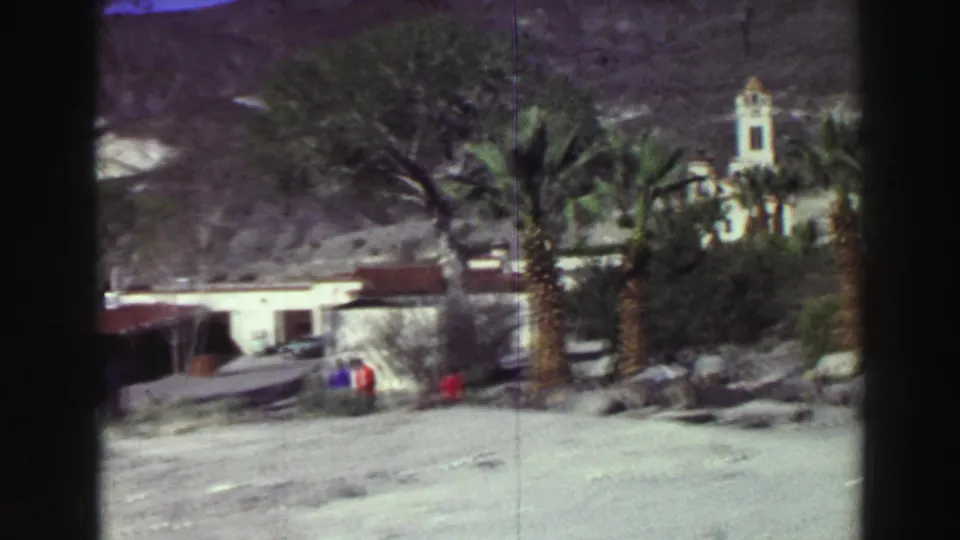 1969 8mm travel view from the Lake Tahoe metadata showing palms, low buildings, rocky open ground, small figures and mountain terrain.