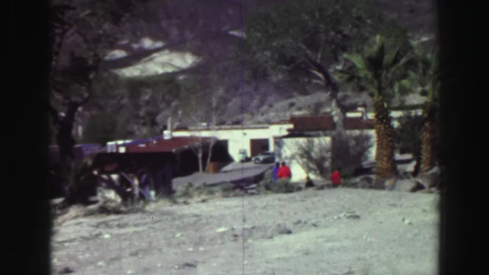 1969 8mm frame from the Lake Tahoe metadata showing a rocky roadside area with palms, low buildings, small figures and mountains.
