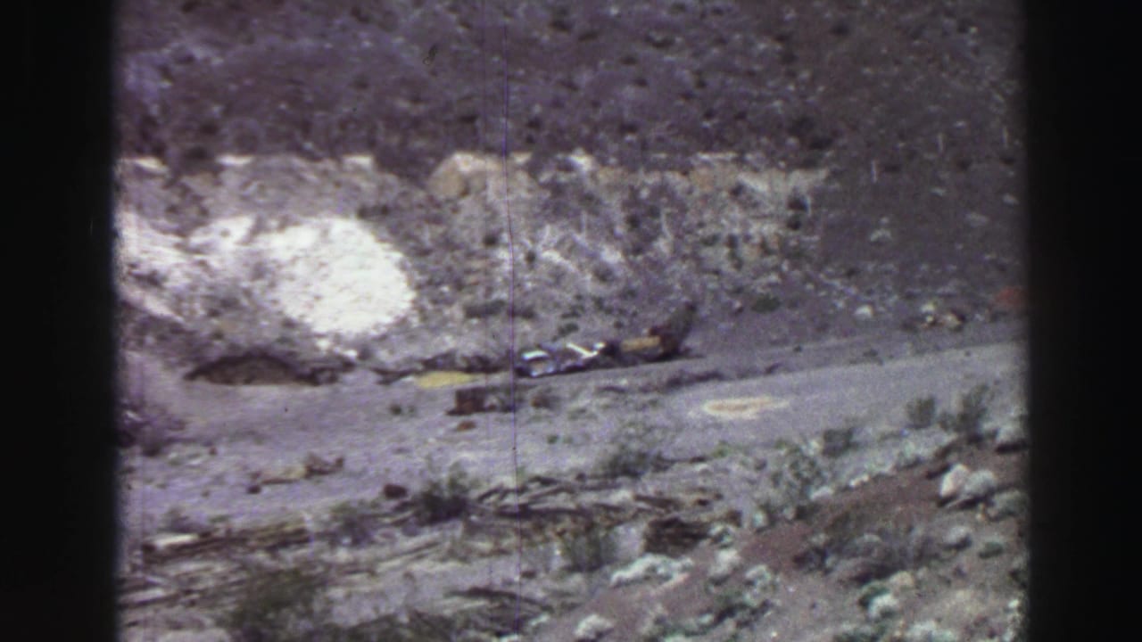 Rocky hillside with a pale patch, dirt track, sparse brush, and scattered debris.