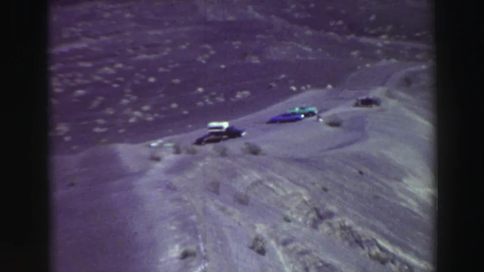 Remote Lake Tahoe-area canyon view with several small cars parked along a dusty ridge road in aged 8mm color.