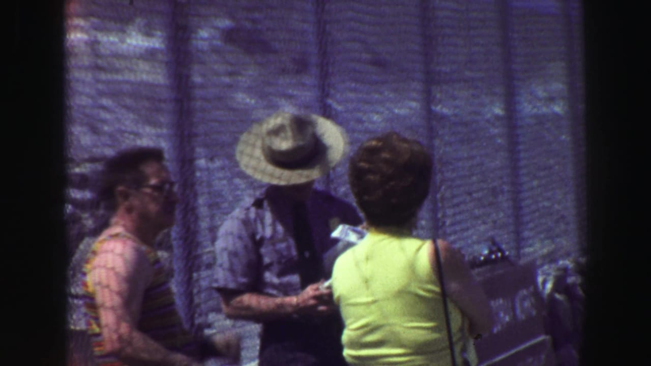 1969 8mm travel footage shows adults at a checkpoint near a chain-link fence as a wide-brimmed uniformed man looks down at documents.