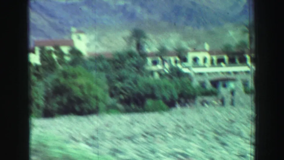 A visitor-shot 8mm view shows a pale building, greenery, palm trees, and dry mountain slopes in an arid landscape.