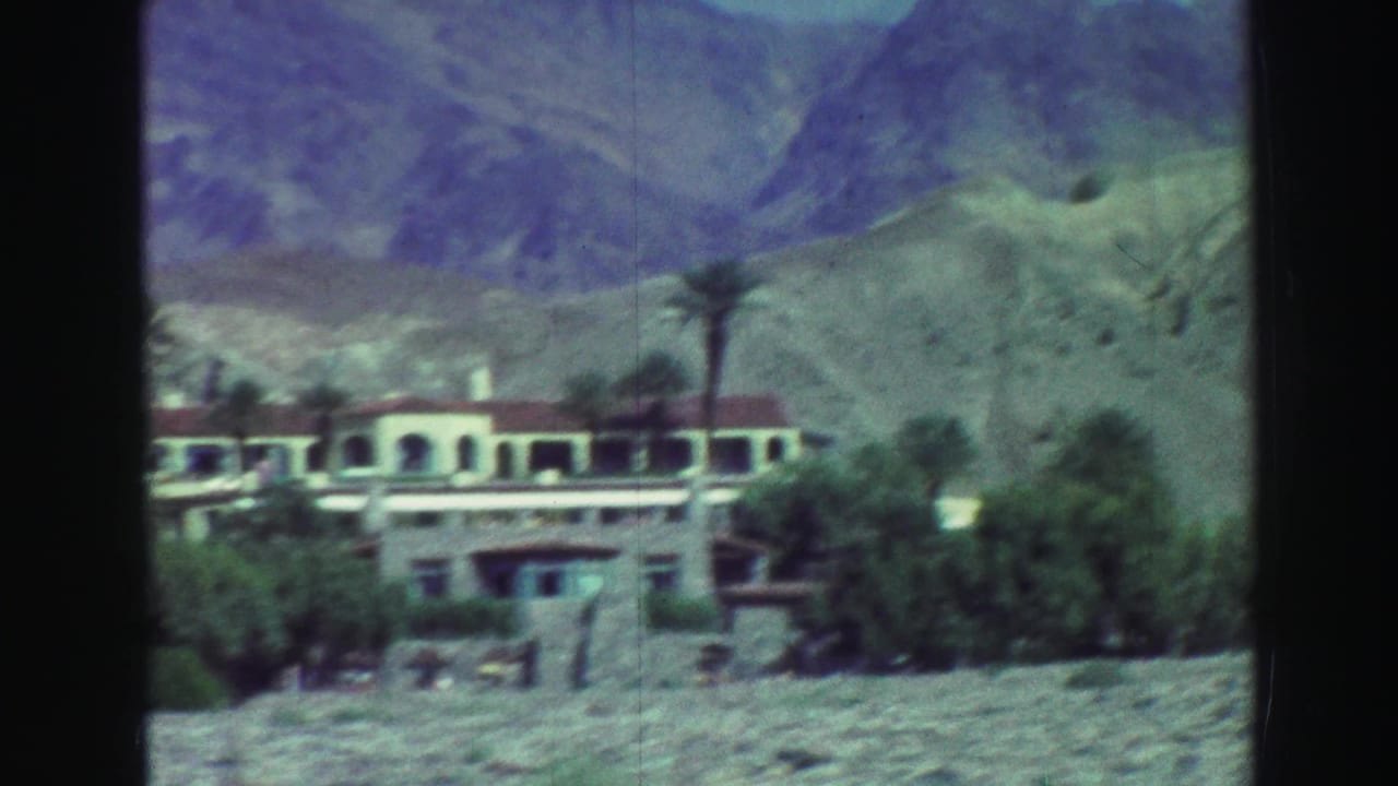 White resort building with red roof, trees, palms, and mountains behind it.