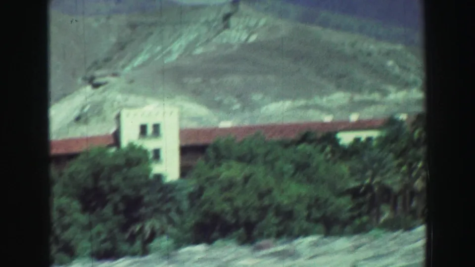 1969 Lake Tahoe 8mm view of a taller white building section with a red-tiled roofline, palms, trees, and pale mountain slopes behind.