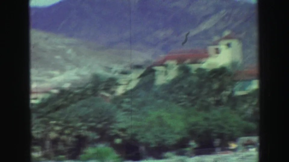 1969 Lake Tahoe travel frame showing a white red-roofed building beyond trees and rocky ground, with blue-gray mountain slopes in the background.
