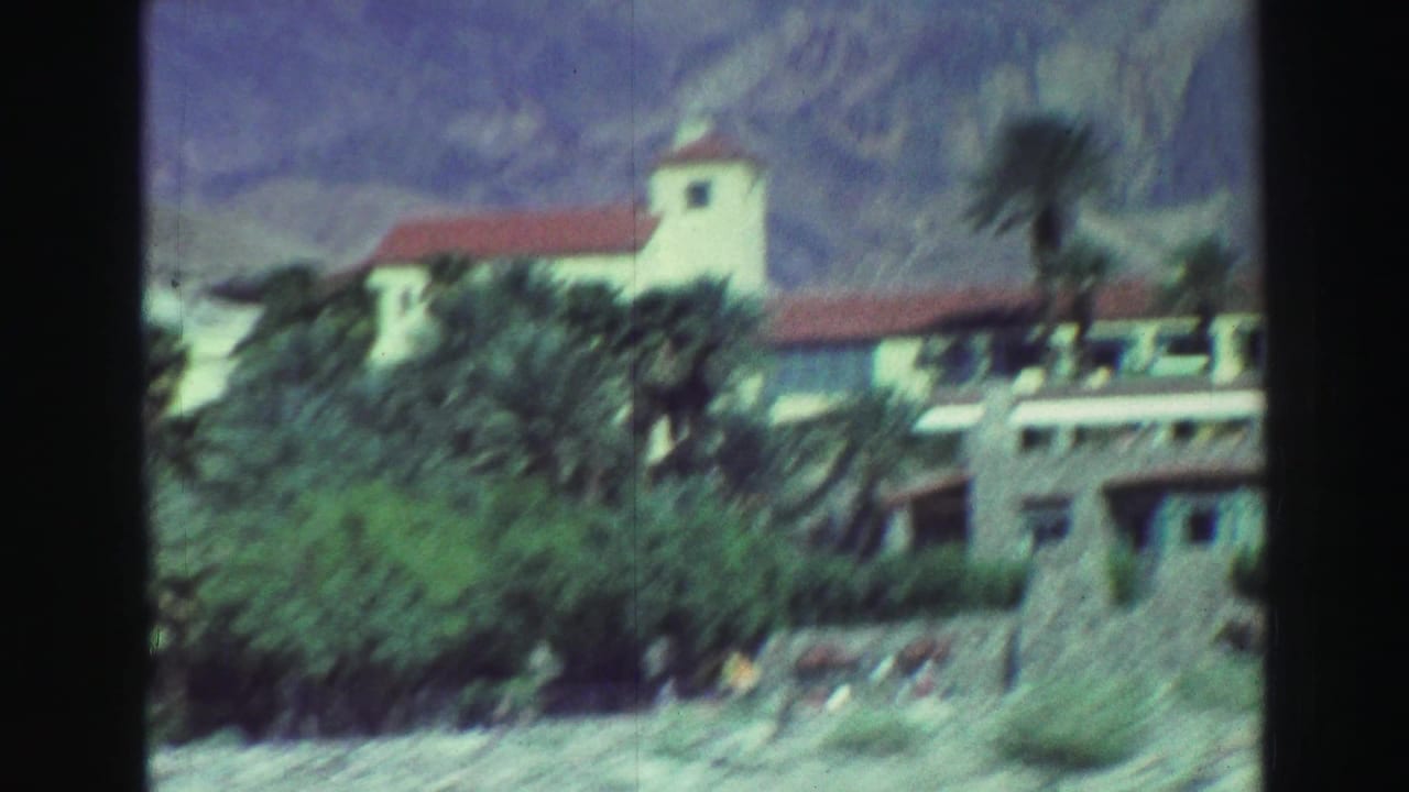 Large white stucco building with red tile roof, palms, shrubs, and mountains behind it.