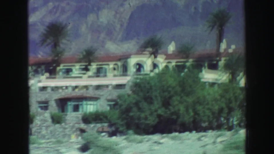 1969 Lake Tahoe-area frame of a Spanish-style exterior with arches, palms, shrubs, and mountain backdrop.