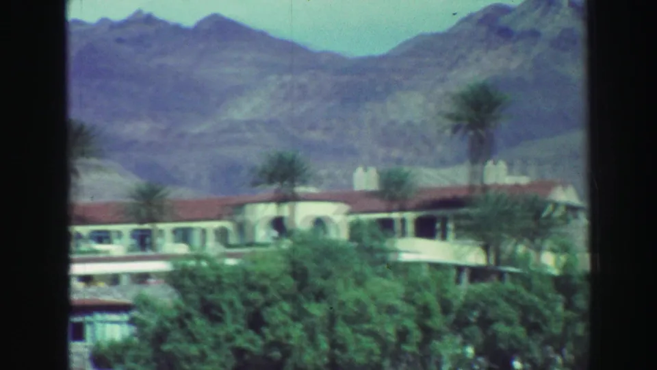 1969 Lake Tahoe-area frame showing a red-roofed stucco building, palms, dense greenery, and rugged mountains.