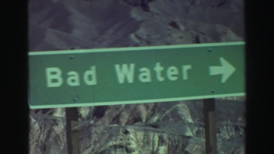 Green roadside sign points toward Bad Water, filmed against rocky desert hills on 8mm home-movie stock.