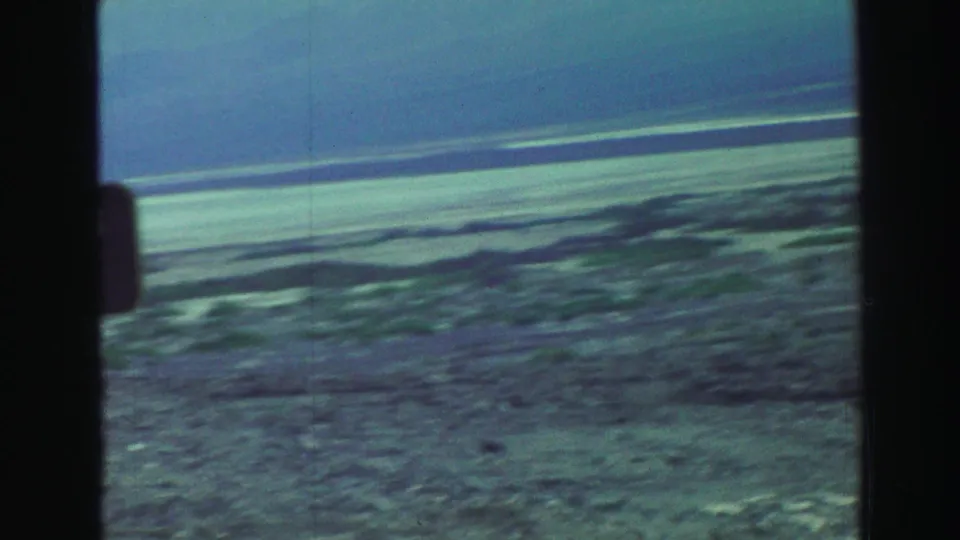 1969 8mm road-trip frame showing a partially visible green sign near open desert scrub and a distant flat horizon.