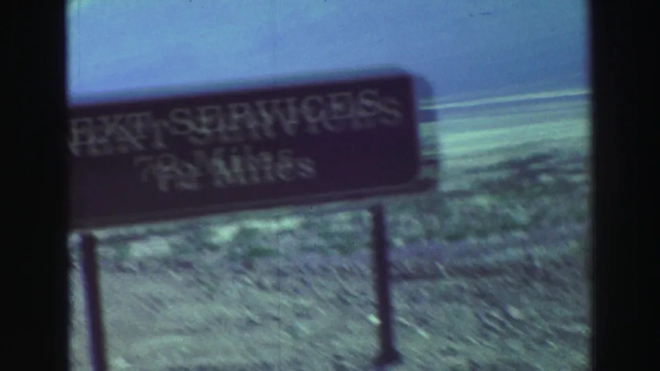 Brown highway service-distance sign stands beside desert terrain, with pale scrubland and a clear sky in the background.
