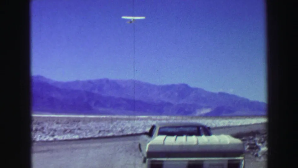 1969 home-movie view of a white glider over a mountain-backed desert scene, with a pale car parked near the road.