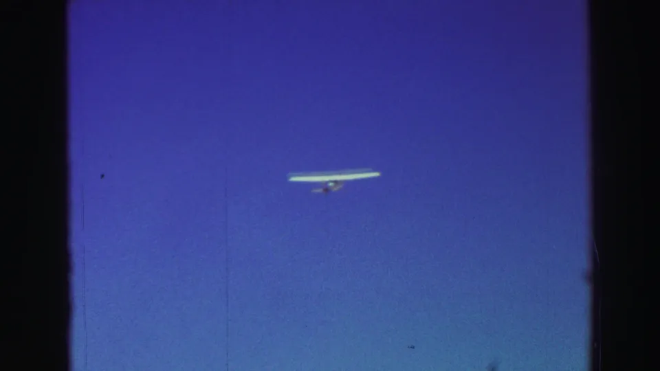 A small white glider appears high in a clear blue sky, framed by dark 8mm film edges and minor scratches.