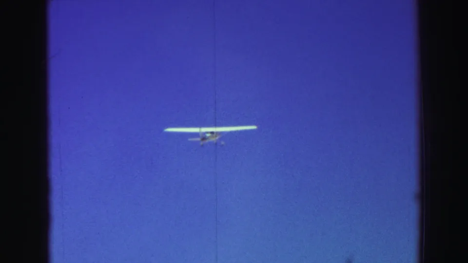 A small aircraft is seen in open blue sky, with visible 8mm grain, vertical marks, and dark frame edges.