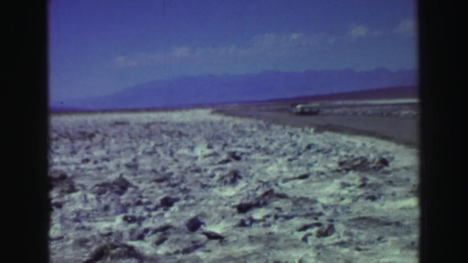 1969 Lake Tahoe-area 8mm frame showing a road through rocky terrain, a distant vehicle, hazy mountains, and blue sky.