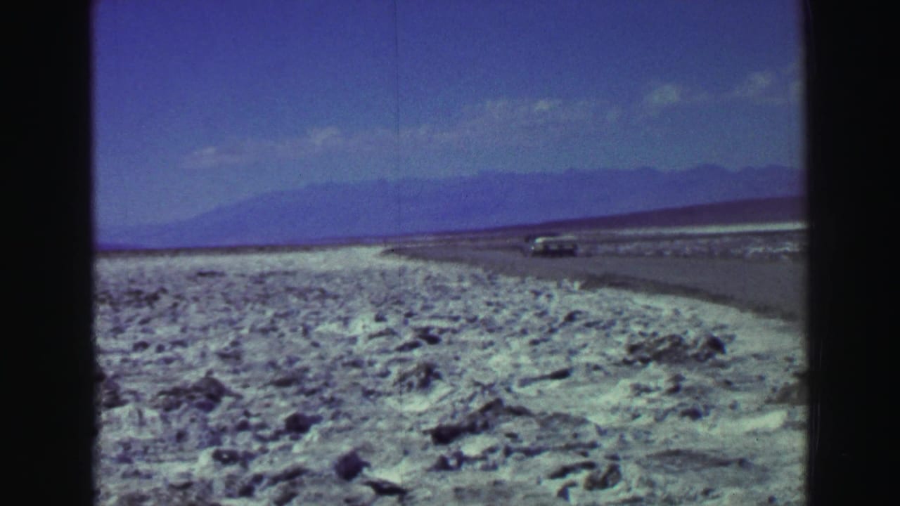 Rocky shoreline with a distant road, car, lake, and mountains under a blue sky.