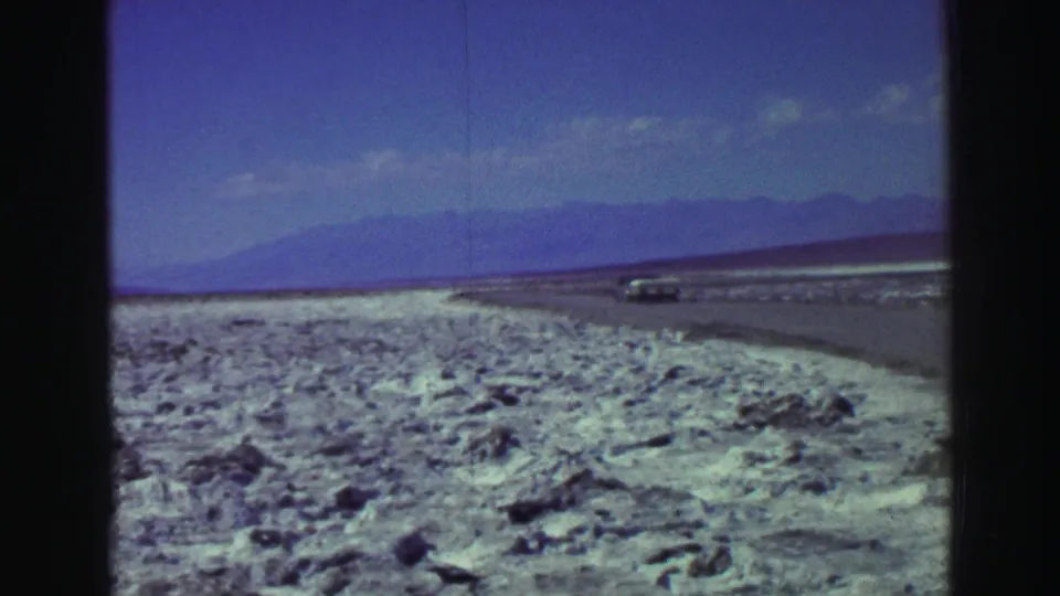 1969 Lake Tahoe home-movie frame showing pale rocky shore, distant water, mountains, and a car on a road at left.