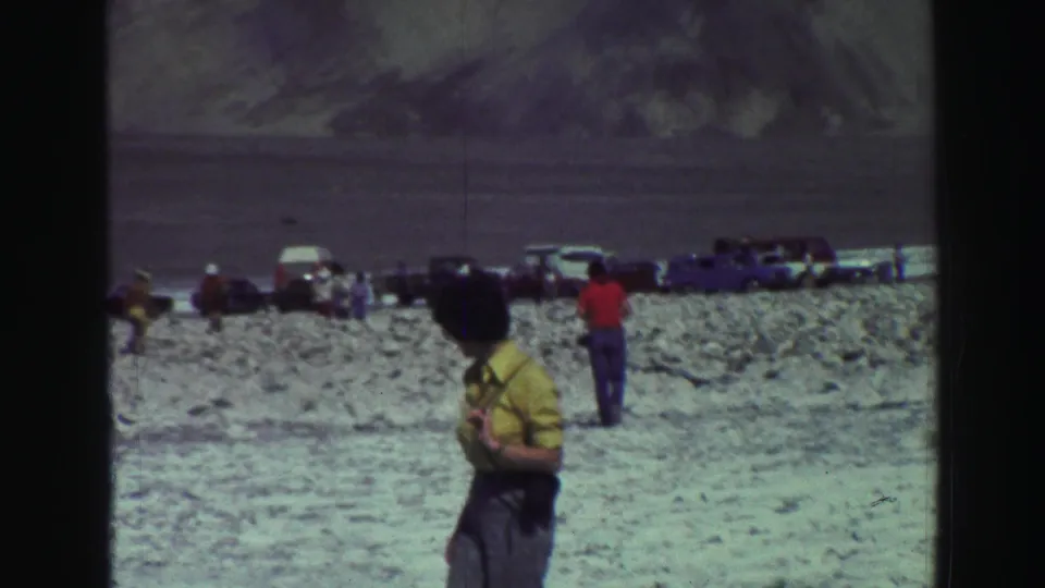 1969 Lake Tahoe frame with visitors near parked vintage cars on pale rocky ground below distant mountains.