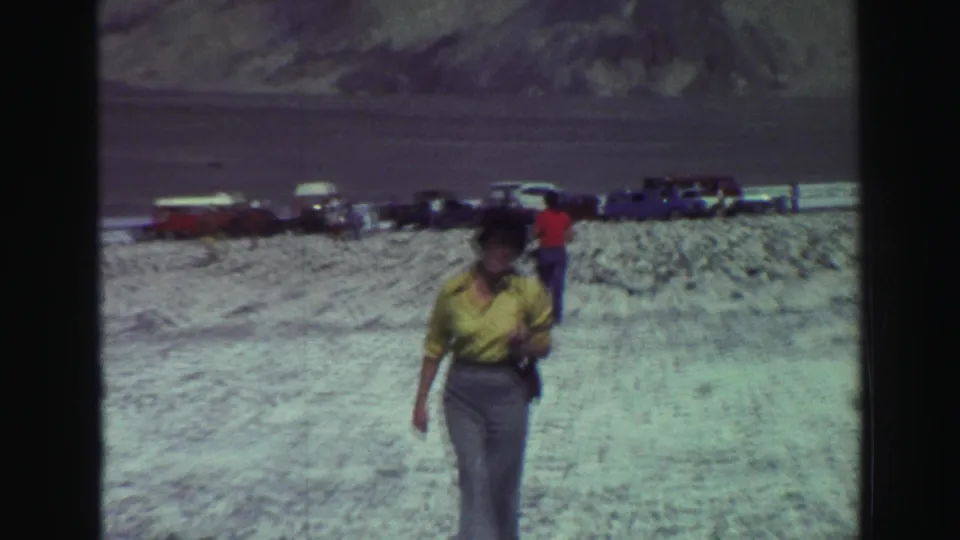 1969 Lake Tahoe frame showing a woman in a yellow shirt walking across rocky terrain with parked cars and visitors behind her.