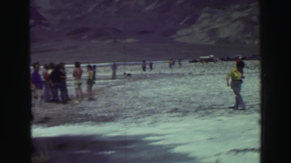 1969 Lake Tahoe frame with a small group near the waterline, mountains beyond, and a woman walking across the rocky shore at right.