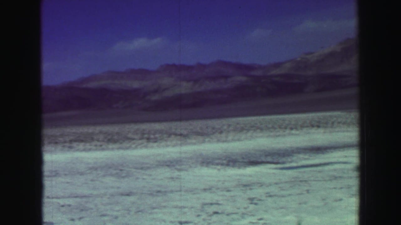 Pale shoreline and distant mountains under a blue sky, seen through dark 8mm frame borders.