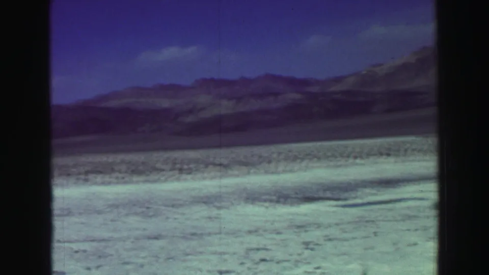1969 Lake Tahoe 8mm frame showing a light beach or shoreline with mountain ridges beyond and a deep blue sky.